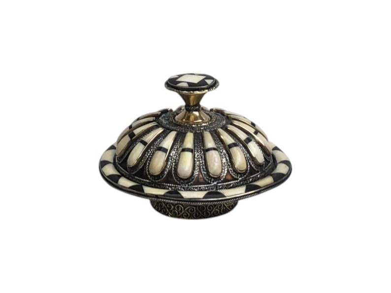 This is a decorative small dish made from a combination of copper and bone. Brass gives it a warm, reflective surface with a hammered texture, while bone inlays add a decorative touch similar to that of ivory, providing a contrast to the metal.