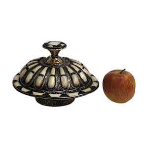This is a decorative small dish made from a combination of copper and bone. Brass gives it a warm, reflective surface with a hammered texture, while bone inlays add a decorative touch similar to that of ivory, providing a contrast to the metal.
