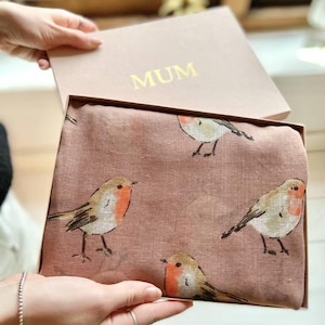 May include: A dusty rose scarf with a robin bird pattern is presented in a matching box. The box lid is open, revealing the scarf with multiple illustrations of the bird. The box is labeled "MUM" in gold lettering.