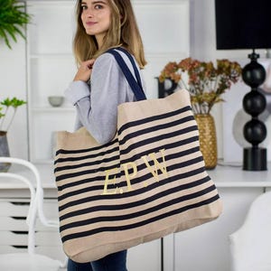 May include: A large beige tote bag with navy blue horizontal stripes. The bag has navy blue straps and gold embroidered initials. The bag is made of a sturdy material and is designed for carrying items.
