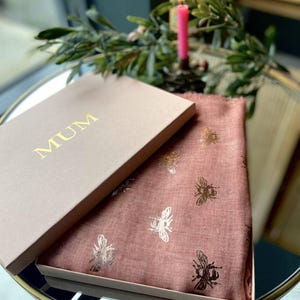 May include: A dusky pink scarf with gold bee motifs is presented in a matching box. The box lid, bearing the gold word "MUM", is open, revealing the scarf. A lit pink candle and foliage are in the background.