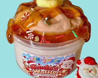 May include: A clear plastic container filled with tan slime topped with amber-colored syrup, a yellow butter pat, and small holiday-themed charms. The label reads Little Loopies Santa's Waffle Scented Buttermilk Pancakes NOT EDIBLE.