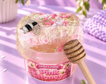 Puede incluir: Un slime transparente y viscoso en un recipiente de plástico con la etiqueta Little Loopies Pink Honey, decorado con un dije de abeja en blanco y negro, chispas de flores rosas y piezas rectangulares blancas, junto a un cucharón de miel de madera.