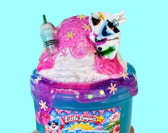 Puede incluir: Un recipiente azul de slime cubierto con slime texturizado blanco y rosa, con un dije de taza y un personaje. La etiqueta dice Little Loopies In Summer Frappé Scented Juniper Twist y Not Edible.