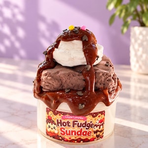 May include: A clear plastic container filled with a textured brown substance resembling a chocolate dessert, topped with white cream, dark brown syrup, and small colorful sprinkles. A label reads Little Loopies Hot Fudge Sundae.