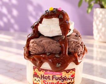 Op de afbeelding: Een doorzichtige plastic container gevuld met een textuurrijke bruine substantie die lijkt op een chocoladedessert, bedekt met witte room, donkerbruine siroop en kleine gekleurde hagelslag. Een label vermeldt Little Loopies Hot Fudge Sundae.
