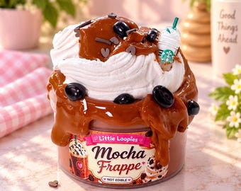 Puede incluir: Un recipiente de plástico transparente lleno de slime color moca cubierto con slime blanco batido, jarabe color caramelo, dijes de granos de café y un dije de vaso de Starbucks en miniatura. La etiqueta dice Little Loopies Mocha Frappe.