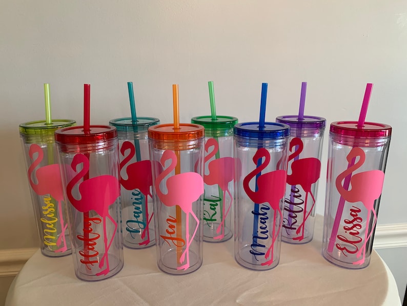 May include: Set of eight clear plastic tumblers with colorful lids and straws. Each tumbler features a pink flamingo silhouette and a personalized name, including "Melissa", "Haley", "Janis", "Jen", "Kat", "Micah", "Kellie", and "Elissa".
