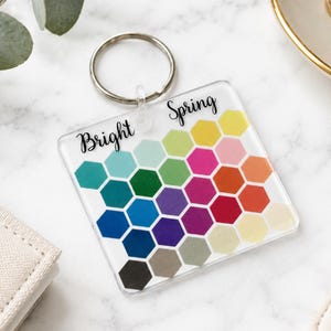 Op de afbeelding: Een vierkante doorzichtige acrylen sleutelhanger met een metalen ring en de tekst Bright Spring in zwarte schrijfletters. Het oppervlak toont een raster van veelkleurige zeshoeken in tinten geel, roze, oranje, groen, blauw, paars, rood, grijs en zwart.