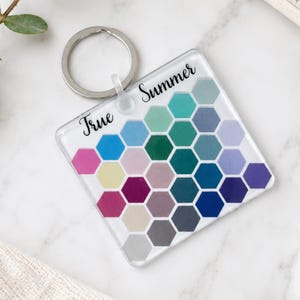 Puede incluir: Un llavero cuadrado de acrílico transparente con una cuadrícula de hexágonos coloridos en varios tonos de azul, verde, morado, rosa y gris. El texto True Summer está impreso en letra cursiva negra en la parte superior del llavero, que incluye una argolla plateada.