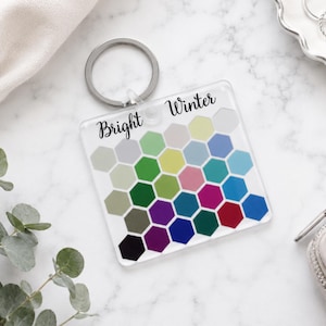 Puede incluir: Un llavero cuadrado de acrílico con una cuadrícula de hexágonos coloridos en varios tonos de verde, azul, rosa, rojo, morado y gris. El texto "Bright Winter" está impreso en letra cursiva negra en la parte superior del llavero con anilla metálica.