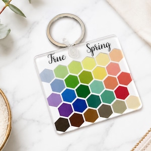 May include: A square clear acrylic keyring featuring a grid of colourful hexagons in various shades including green, blue, yellow, red, and brown. The text True Spring is printed in black script at the top of the keyring, which includes a silver metal ring.