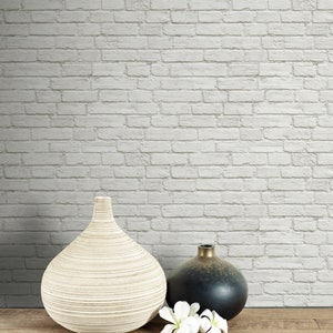 May include: A large, light-coloured, ribbed ceramic vase sits next to a smaller, round, dark-toned vase with a metallic finish. Two white flower blossoms are placed in front of the dark vase, all resting on a flat, horizontal wooden surface.