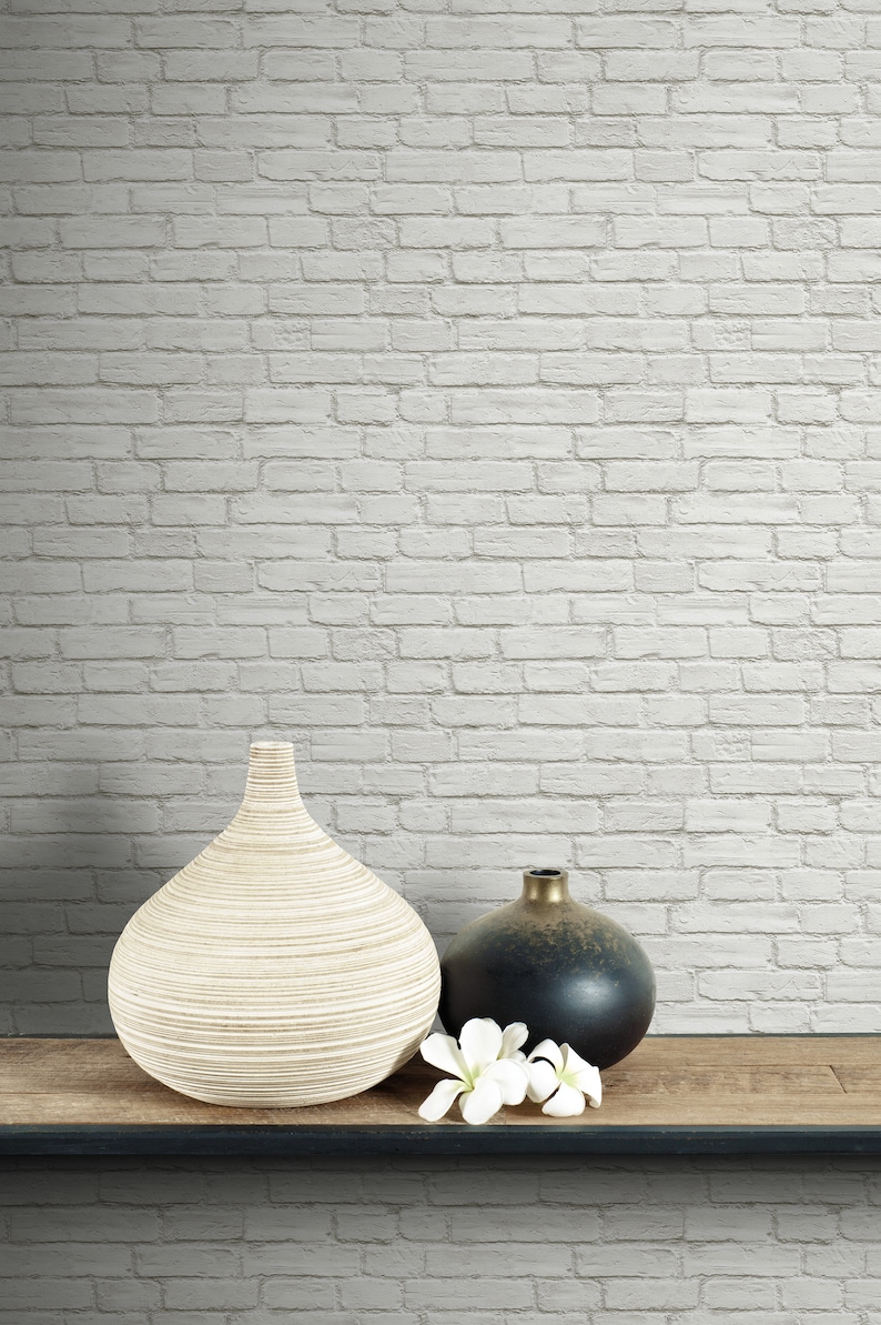May include: A large, light-coloured, ribbed ceramic vase sits next to a smaller, round, dark-toned vase with a metallic finish. Two white flower blossoms are placed in front of the dark vase, all resting on a flat, horizontal wooden surface.