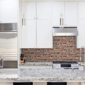 May include: A kitchen interior featuring white flat-panel cabinetry, a stainless steel refrigerator, and a gas range. The backsplash is made of red and brown brick, and the countertop is a speckled grey and white granite with a metal faucet.