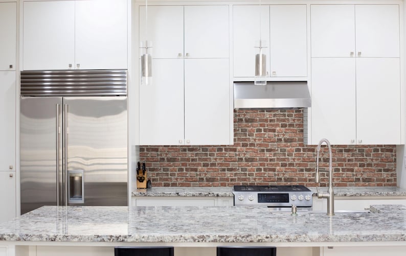 May include: A kitchen interior featuring white flat-panel cabinetry, a stainless steel refrigerator, and a gas range. The backsplash is made of red and brown brick, and the countertop is a speckled grey and white granite with a metal faucet.