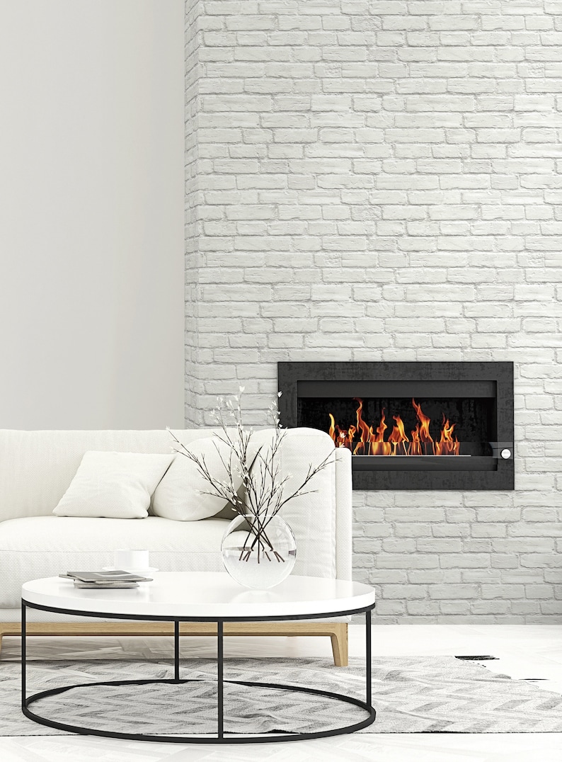 May include: A rectangular black fireplace insert with visible orange and yellow flames is mounted on a white brick wall. A white sofa with cushions sits in front of the fireplace, accompanied by a round white coffee table with a glass vase.