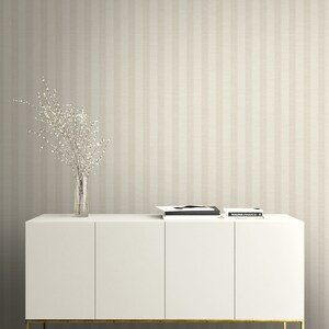 K&ouml;nnte beinhalten: Ein wei&szlig;es Sideboard mit vier T&uuml;ren und einem goldfarbenen Metallgestell, darauf eine klare Glasvase mit wei&szlig; bl&uuml;henden Zweigen und ein Stapel B&uuml;cher. Der Hintergrund zeigt eine Tapete mit vertikalen Streifen in Beige und Creme.