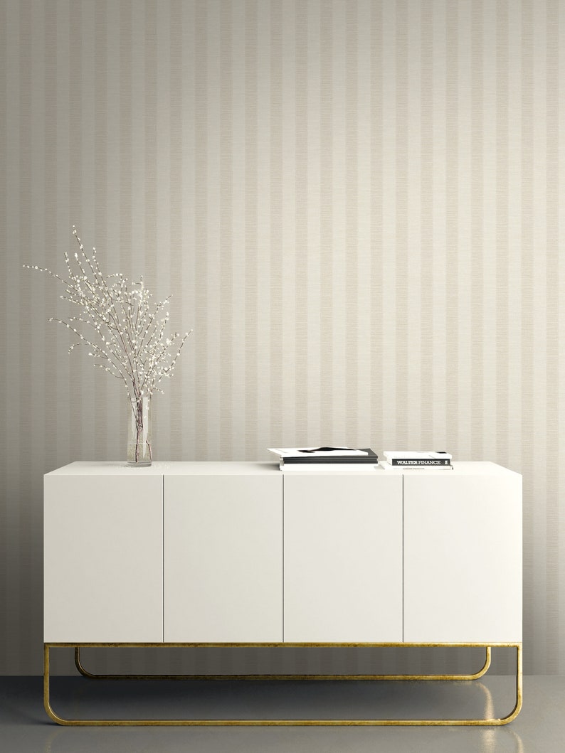 K&ouml;nnte beinhalten: Ein wei&szlig;es Sideboard mit vier T&uuml;ren und einem goldfarbenen Metallgestell, darauf eine klare Glasvase mit wei&szlig; bl&uuml;henden Zweigen und ein Stapel B&uuml;cher. Der Hintergrund zeigt eine Tapete mit vertikalen Streifen in Beige und Creme.