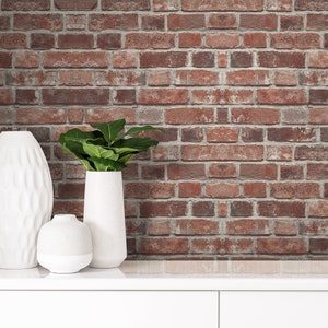May include: Three white ceramic vases of varying shapes and sizes, with one containing a green leafy plant, positioned in front of a wall featuring a red and brown brick pattern with visible grey mortar lines between the rectangular bricks.
