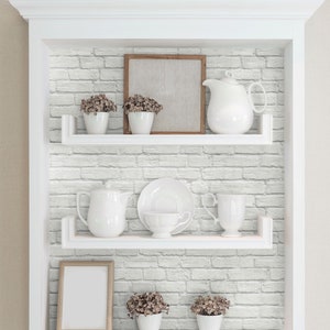 May include: A white built-in shelving unit with three shelves against a white faux brick wall. The shelves hold white ceramic pitchers, cups, saucers, small potted plants with brown flowers, and two empty wooden picture frames for display.