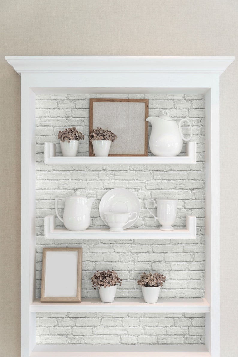 May include: A white built-in shelving unit with three shelves against a white faux brick wall. The shelves hold white ceramic pitchers, cups, saucers, small potted plants with brown flowers, and two empty wooden picture frames for display.