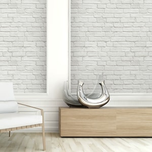 May include: Two decorative vases with a crescent moon shape, one crafted from clear glass and the other featuring a polished silver metallic finish, positioned on a light wood console table against a white brick wall with a white chair nearby.