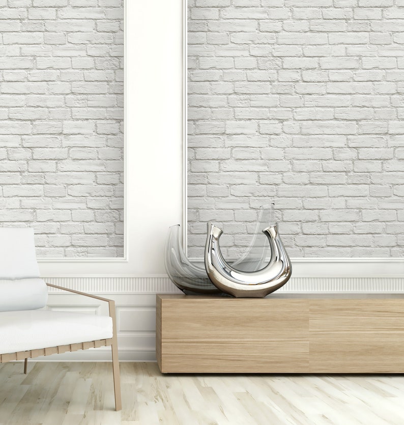 May include: Two decorative vases with a crescent moon shape, one crafted from clear glass and the other featuring a polished silver metallic finish, positioned on a light wood console table against a white brick wall with a white chair nearby.