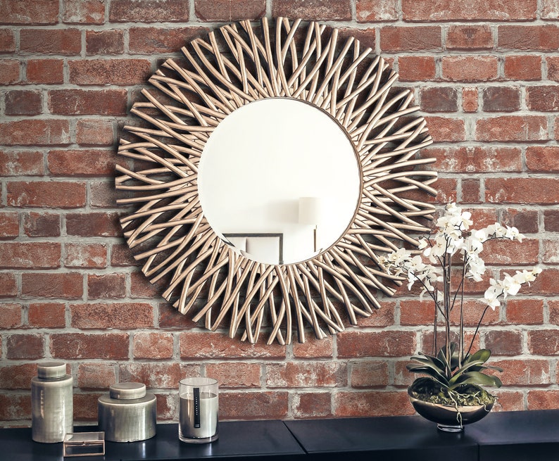 May include: A round wall mirror featuring a decorative frame composed of numerous thin, curved metallic rods arranged in a sunburst pattern. The mirror has a reflective glass centre and is mounted on a wall with a textured red brick surface.