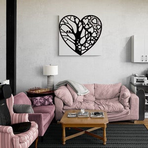 May include: A black metal wall art sculpture in the shape of a heart with a tree and birds on one side and a pattern of hearts on the other side. The art is hanging on a white wall above a pink sofa and a coffee table.