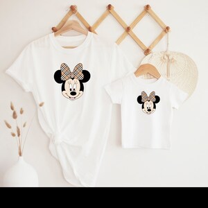 May include: Two white t-shirts featuring a cartoon image of Minnie Mouse wearing a brown and black checked bow. The larger shirt is folded and the smaller shirt is laid flat.