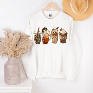 May include: White sweatshirt with a graphic of four different coffee drinks. The drinks include a pumpkin spice latte, a caramel macchiato, a peppermint mocha, and a regular coffee.