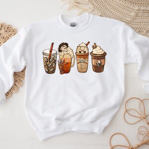 May include: White sweatshirt with a graphic of four different coffee drinks. The drinks are a pumpkin spice latte, a caramel macchiato, a mocha, and a regular coffee. Each drink has a spooky Halloween character on it.