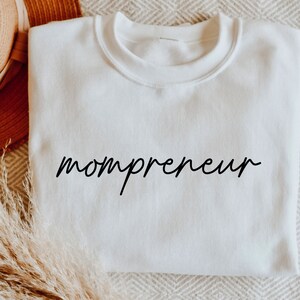 May include: A white crew neck sweatshirt with the word "mompreneur" in black script. The sweatshirt is made of a soft material and is displayed on a neutral-toned surface. The overall aesthetic is simple and modern.