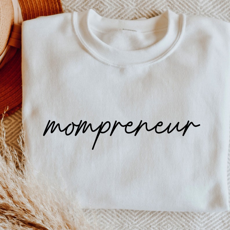 May include: A white crew neck sweatshirt with the word "mompreneur" in black script. The sweatshirt is made of a soft material and is displayed on a neutral-toned surface. The overall aesthetic is simple and modern.