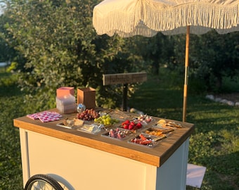 May include: A white wooden cart with a wooden top and a silver wheel. The cart is filled with various snacks, including fruit, cheese, and crackers. The cart is set up outdoors under a white fringed umbrella.