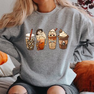 May include: A gray sweatshirt with a graphic of four different coffee drinks. The drinks are decorated with Halloween-themed characters, including a ghost, a pumpkin, and a vampire.