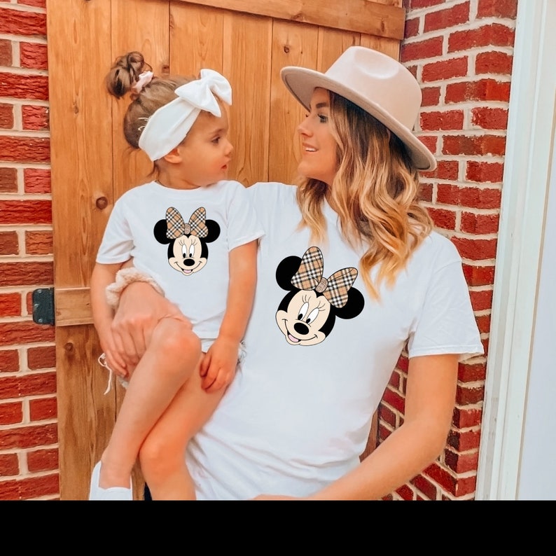 May include: A white t-shirt featuring a cartoon image of Minnie Mouse wearing a plaid bow. The shirt is being worn by a woman and a young girl. The woman is wearing a beige hat.