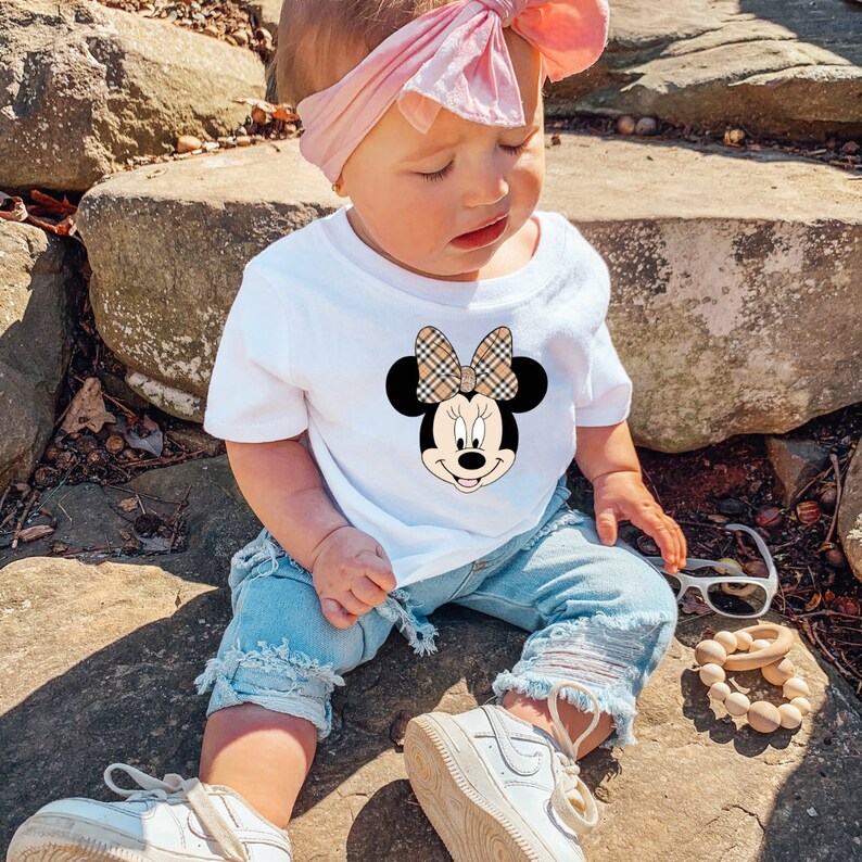 May include: A white t-shirt featuring a cartoon image of Minnie Mouse wearing a plaid bow. The shirt is being worn by a young child sitting on rocks wearing blue denim shorts and white trainers.