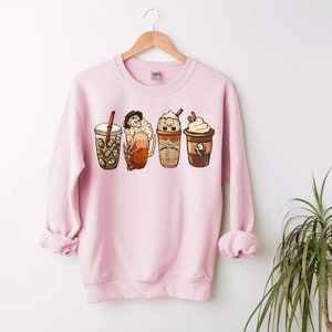May include: Pink sweatshirt with a graphic of four different coffee drinks with whipped cream and toppings. The drinks are in a row from left to right.
