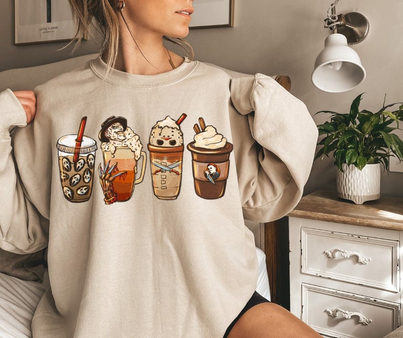 May include: A beige sweatshirt with a graphic print of four different coffee drinks. The drinks are decorated with whipped cream, chocolate syrup, and a character from a horror movie.