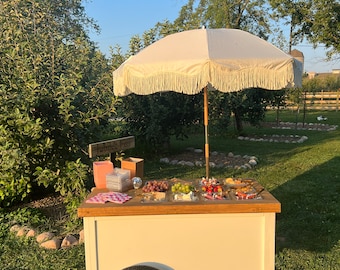 Op de afbeelding: Een witte houten kar met een houten bovenkant en een witte parasol. De kar is gevuld met verschillende snacks en lekkernijen, waaronder druiven, aardbeien, kaas en crackers. De kar is gelabeld met "Bubbly Fun" en staat op een grasveld.