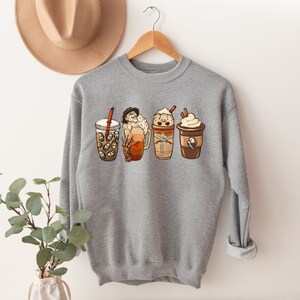 May include: Gray sweatshirt with a graphic of four different coffee drinks with whipped cream and toppings. The drinks are arranged in a row from left to right.