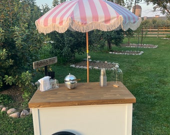 May include: A white and wood ice cream cart with a pink and white striped umbrella. The cart has a wooden countertop, a single wheel, and a disco ball. The sign reads "Jubilee Fuji".