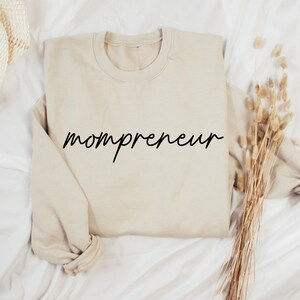 May include: A beige sweatshirt with the word "mompreneur" in black cursive script across the front. The sweatshirt is folded on a white surface, with dried ornamental grass to the right.