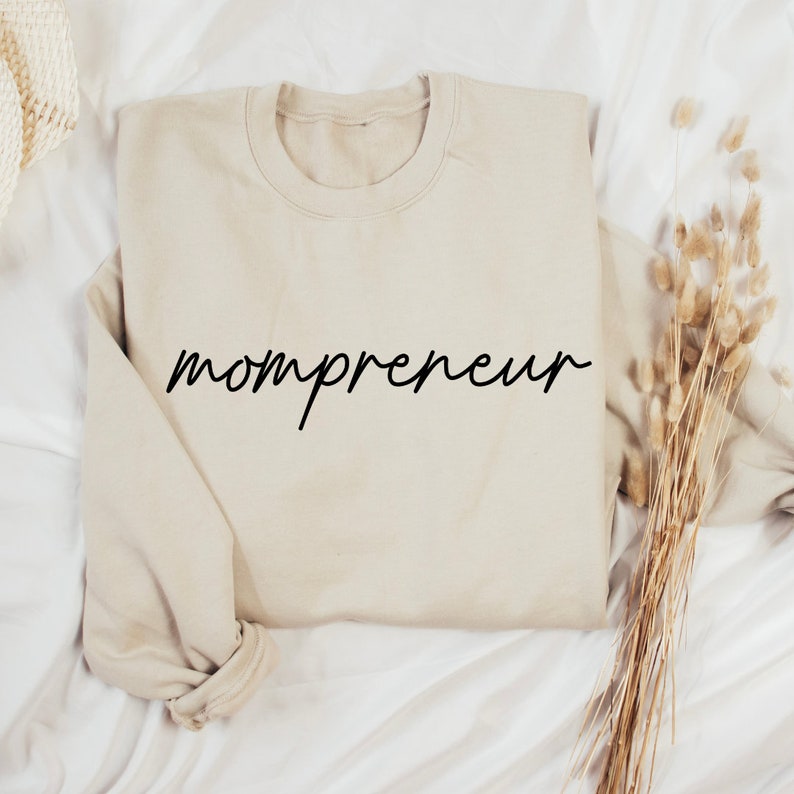 May include: A beige sweatshirt with the word "mompreneur" in black cursive script across the front. The sweatshirt is folded on a white surface, with dried ornamental grass to the right.