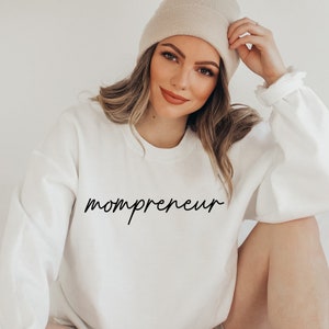 May include: A white sweatshirt with the word "mompreneur" in black script. The sweatshirt has long sleeves and a crew neck. A woman is wearing the sweatshirt and a beige beanie.