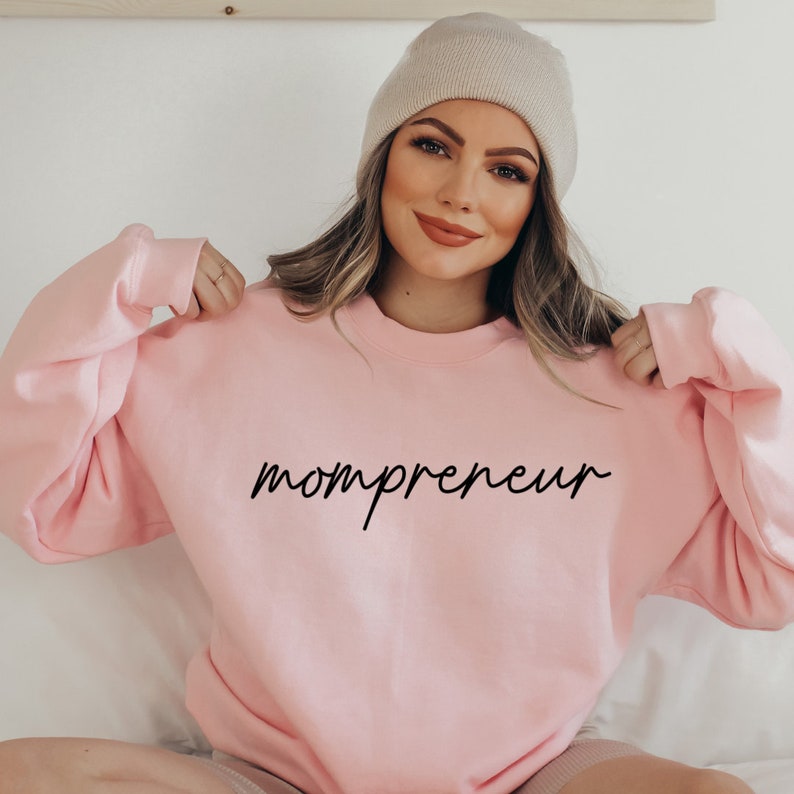 May include: A pale pink crew neck sweatshirt with the word "mompreneur" in black script. The model is wearing a beige beanie. The sweatshirt is a casual style.