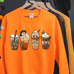 May include: Orange sweatshirt with a graphic of four Halloween-themed coffee drinks. The drinks feature characters from horror movies, including Freddy Krueger, Jason Voorhees, and Michael Myers.