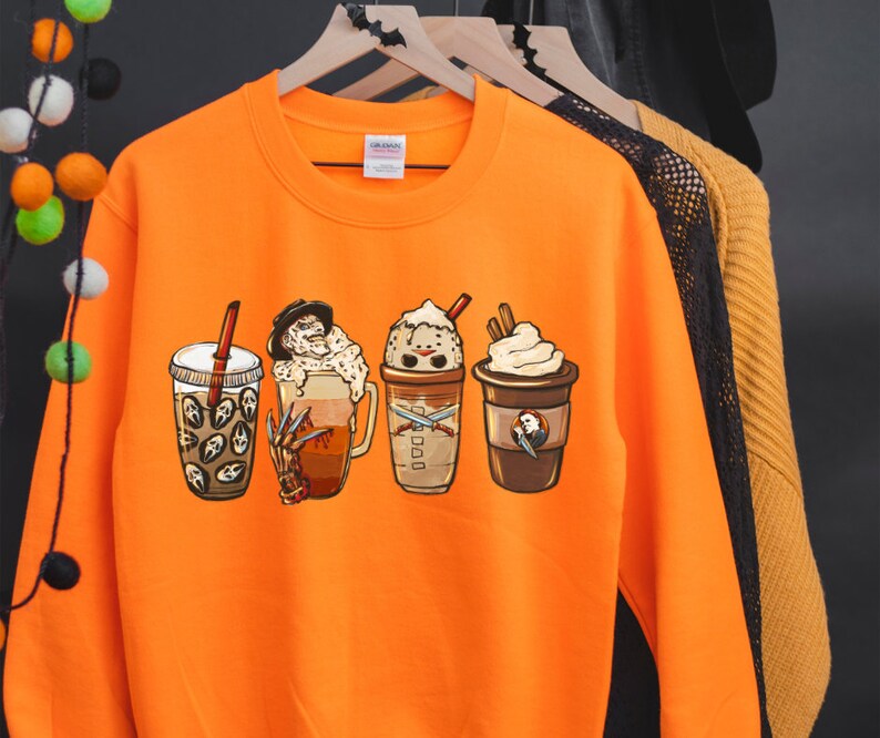 May include: Orange sweatshirt with a graphic of four Halloween-themed coffee drinks. The drinks feature characters from horror movies, including Freddy Krueger, Jason Voorhees, and Michael Myers.